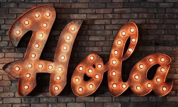 A large illuminated “Hola” sign in bulb-lit script letters on a brick wall