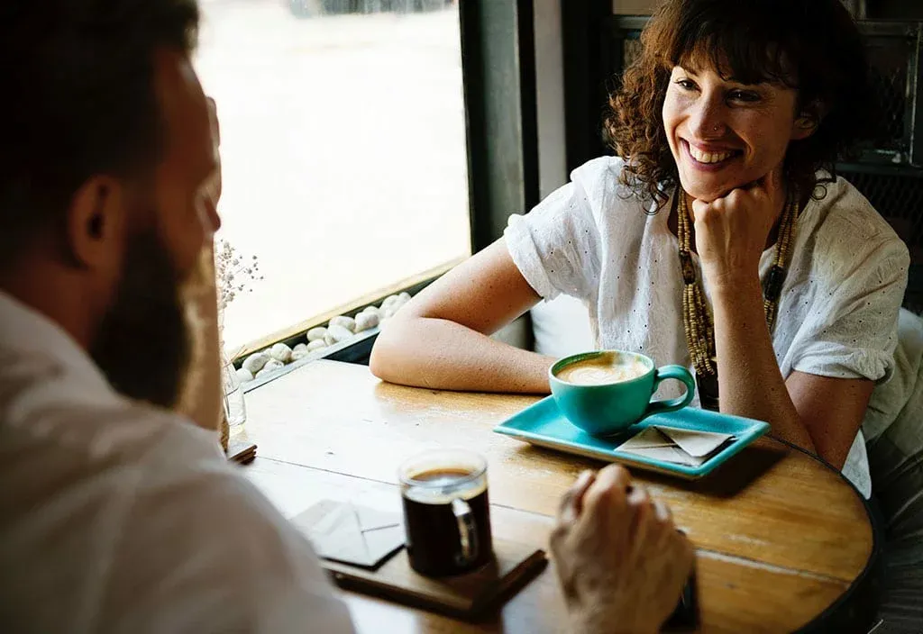 A smiling woman chats with a bearded man over coffee at a wooden cafe table