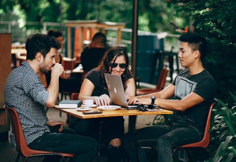 Three friends at an outdoor cafe table, one typing on a laptop as others look on