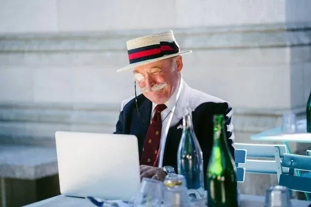 An older man in a straw boater and monocle smiling at a laptop outside a cafe